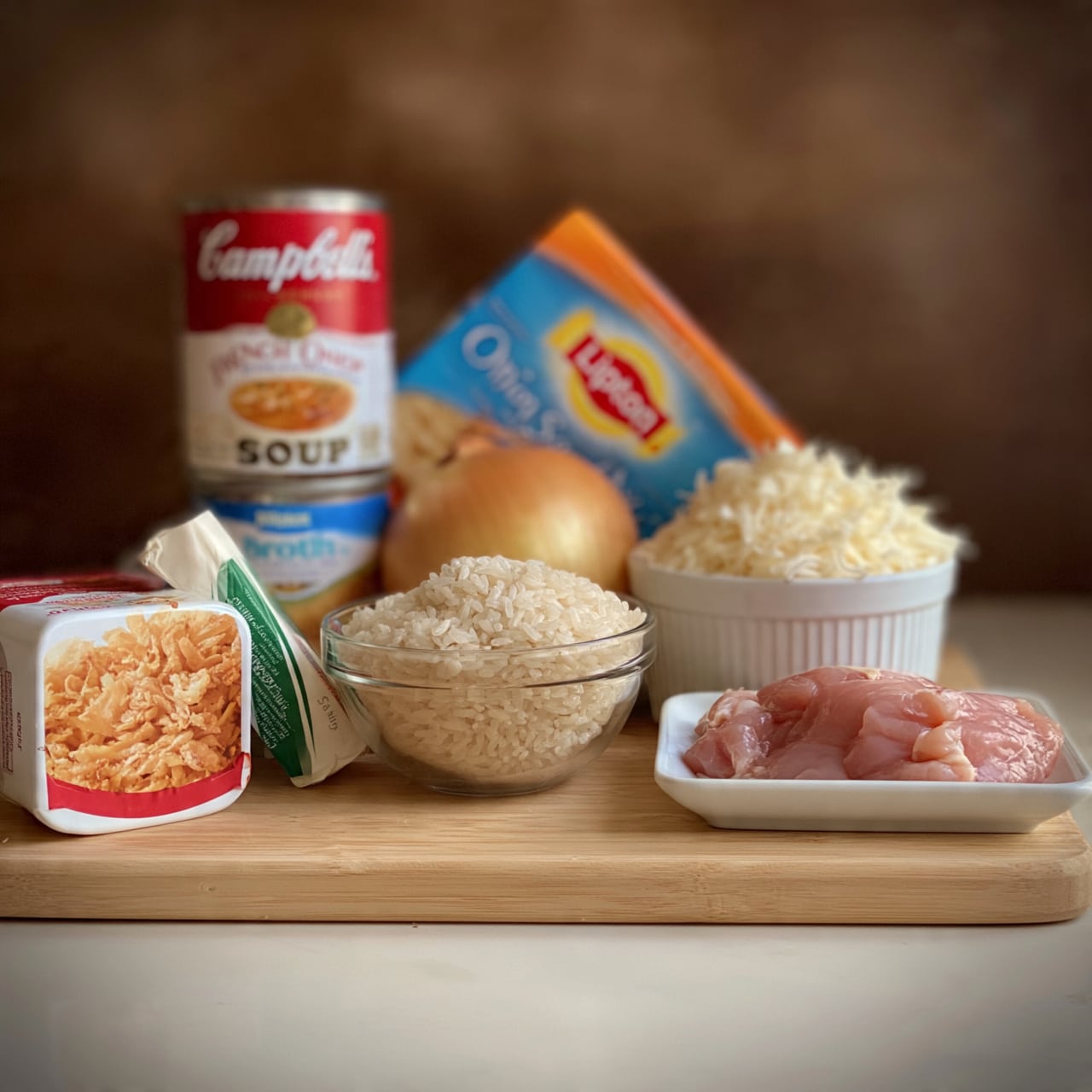 square image A collection of ingredients is arranged on a light wooden board with a brown blurred background. On the left, there is a white and red container of crispy fried onions with a green and white stick of salted butter leaning on it. Behind these, a red and white can of Campbell’s French onion soup sits on top of a blue and white can of chicken broth. Next, a large yellow onion is placed in front of a clear glass bowl filled with uncooked white rice. Behind the rice bowl, a blue and orange box of Lipton onion soup mix leans slightly back. To the right, a white ceramic ramekin is piled high with shredded white cheese, and next to it, raw pink chicken pieces rest on a small white rectangular dish. photo taken with an iphone --ar 4:5 --v 7