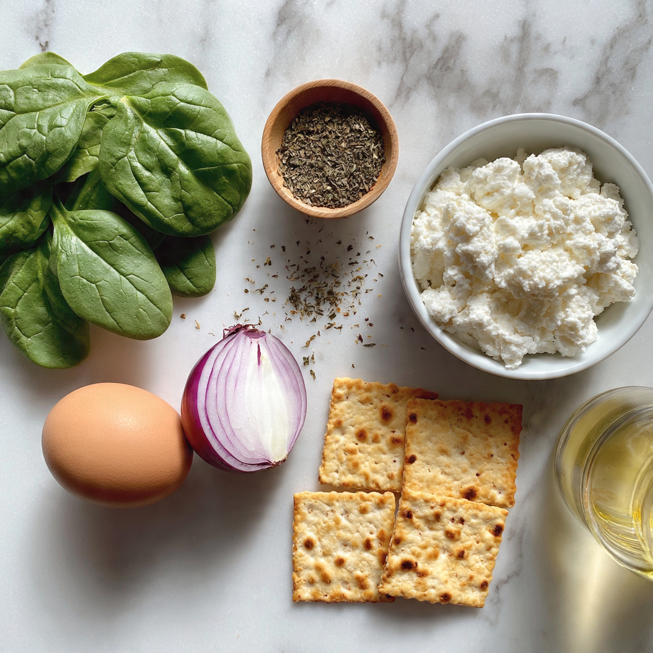 square image The image shows a white marbled surface with fresh ingredients neatly arranged. On the left side, there is a group of bright green spinach leaves with smooth textures. Below the spinach is a whole brown egg and next to it is half of a red onion with purple skin and white inside. Moving right, there is a small wooden bowl filled with dried herbs, and some loose herbs scattered nearby. A white bowl holds fluffy white cottage cheese. Below the bowl are four golden-brown, square crackers with small toasted spots. On the far right is a glass container with light yellow olive oil. Photo taken with an iphone --ar 4:5 --v 7