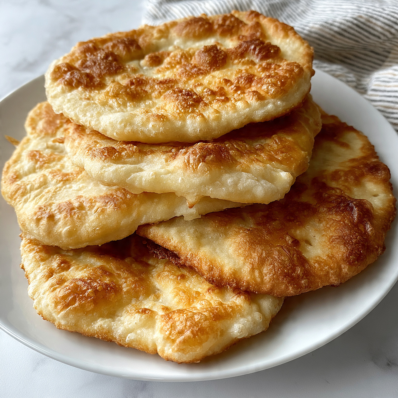 square image The image shows a white round plate with five pieces of fried flatbread stacked unevenly. Each flatbread has a golden brown and light tan color with bubbly, crispy texture on top. The flatbreads are slightly puffy with some parts looking smooth and others showing rough, flaky patches. The plate is placed on a white marbled surface with a striped cloth partially visible in the background. Photo taken with an iphone --ar 4:5 --v 7