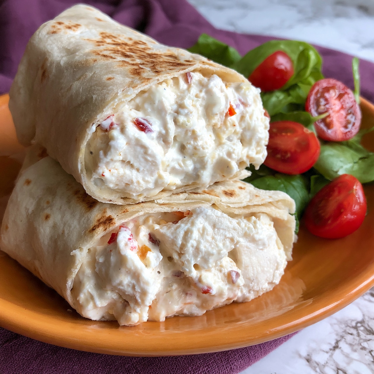 square image A close-up of a tortilla wrap cut in half on an orange plate, showing two layers inside: the first layer is creamy white sauce mixed with chunks of white chicken pieces and small red chili bits, the second layer is the soft, slightly toasted beige tortilla wrap holding the filling together; next to the wrap is a small side salad with fresh green leaves and halved cherry tomatoes, all placed on a purple cloth with a white marbled surface underneath. photo taken with an iphone --ar 4:5 --v 7