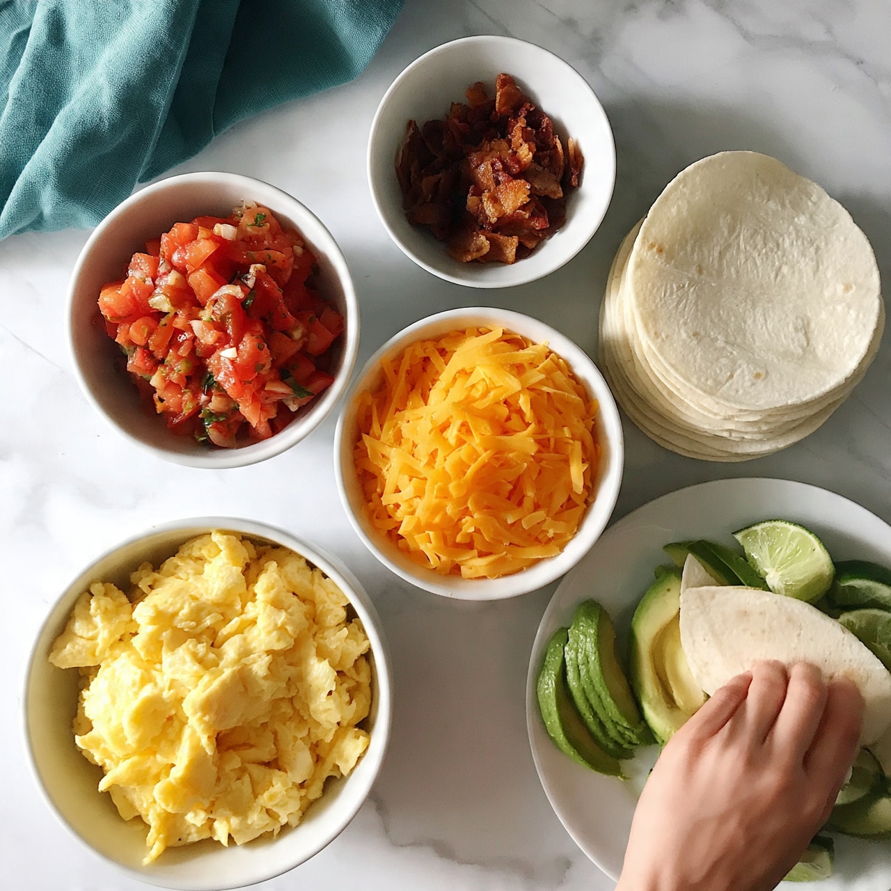 square image The image shows six white bowls and plates arranged neatly on a white marbled surface. Starting from the bottom left, there is a bowl filled with fluffy light yellow scrambled eggs. Above it, a bowl contains a fresh red salsa made of chopped tomatoes, onions, and herbs. To the right of that, a bowl is full of bright orange shredded cheddar cheese. Above the cheese, a smaller bowl holds bits of cooked brown bacon. To the right of the bacon bowl, there is a stack of plain white tortillas. Below the tortillas, a white plate holds slices of fresh green avocado with lime wedges on the side. A woman's hand is about to pick a tortilla. There is a folded teal cloth napkin near the top left corner of the image photo taken with an iphone --ar 4:5 --v 7