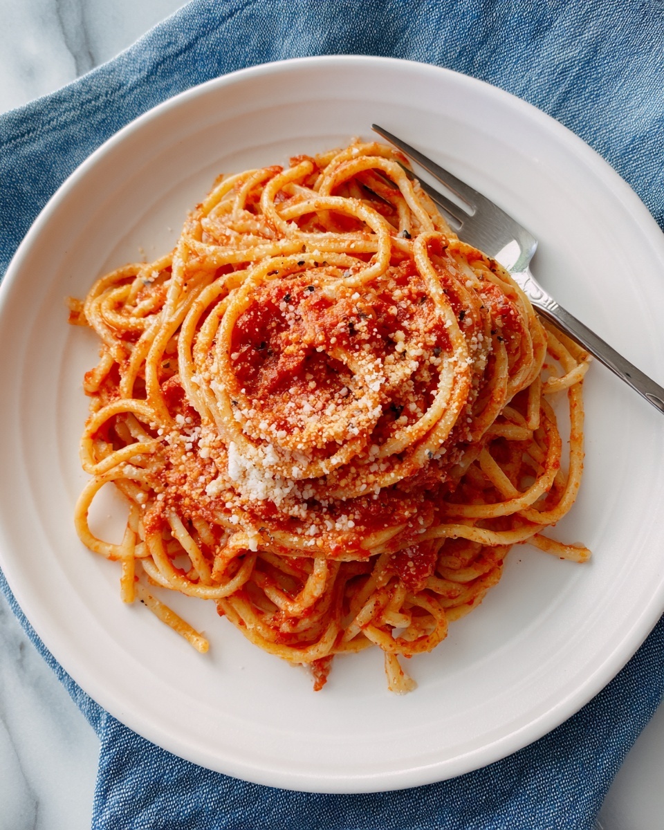 A white plate holds a serving of spaghetti pasta mixed with red tomato sauce, topped with a sprinkle of grated cheese that looks slightly coarse and white. The spaghetti strands are twisted together, and there is a silver fork resting on the right side of the plate. The plate is placed on a blue cloth with a white marbled surface underneath. photo taken with an iphone --ar 4:5 --v 7
