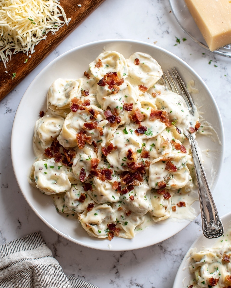 A plate filled with creamy white tortellini pasta covered in a smooth, thick white sauce. The tortellini are layered in a single pile with bits of crispy brown bacon scattered on top and mixed inside. Small green herb pieces are sprinkled throughout the dish, adding a touch of color. The plate is plain white and set on a white marbled surface. A silver fork rests on the edge of the plate, slightly cleaning the sauce. Nearby, there is a wooden board with shredded cheese and a glimpse of another white plate with more tortellini. photo taken with an iphone --ar 4:5 --v 7