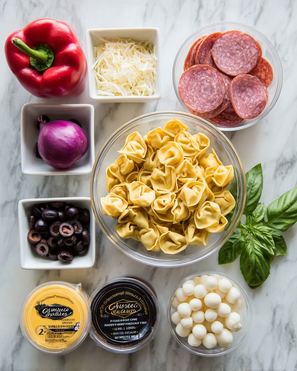 A clear glass bowl filled with yellow tortellini sits surrounded by small white bowls containing grated cheese, dark brown dressing, sliced black olives, and round slices of salami. Near the bowls are a bright red bell pepper, a small red onion, a container of fresh mozzarella pearls, and a yellow container of red cherry tomatoes labeled