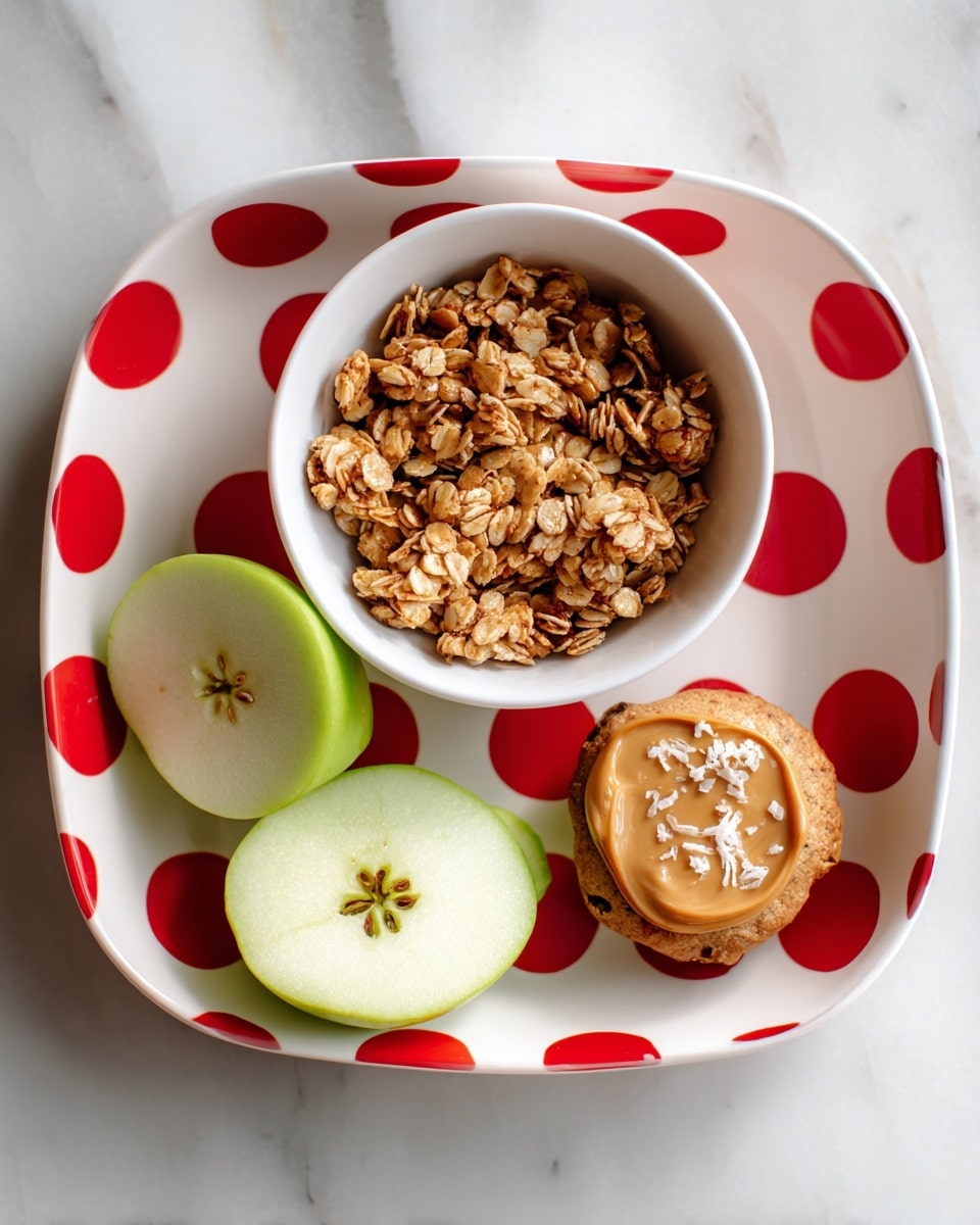 A white square plate with large red polka dots holds three items on a white marbled surface: a white bowl filled with crunchy granola containing oats and nuts on the left, two halves of a green apple showing the light green skin and smooth pale inside with star-shaped core seeds in the center and bottom part of the plate, and a cookie topped with peanut butter and sprinkled with shredded coconut resting partially underneath the apple halves on the right side. Photo taken with an iphone --ar 4:5 --v 7