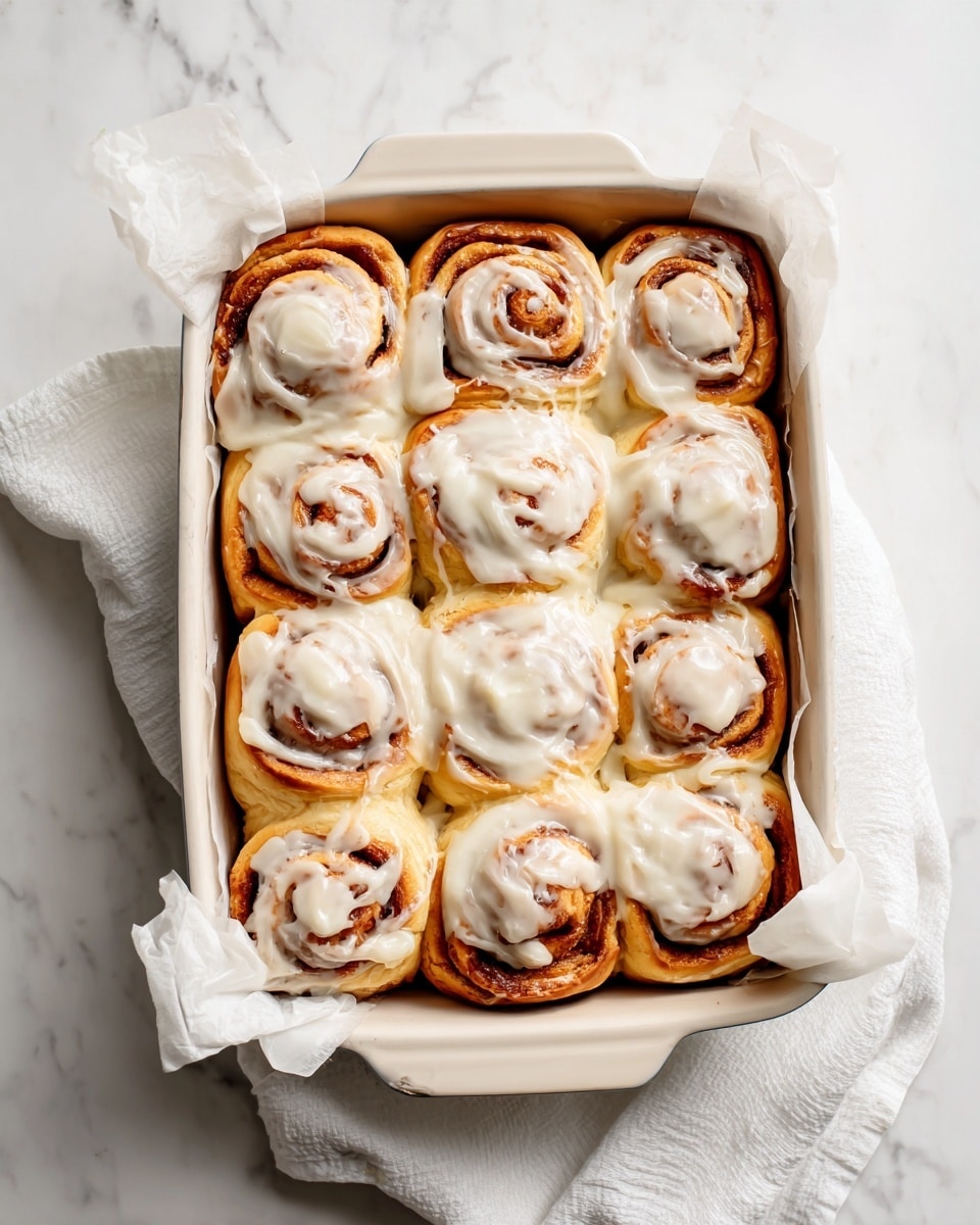 A white rectangular baking dish filled with twelve cinnamon rolls tightly arranged in three rows of four. Each roll is a light golden brown with visible spirals of cinnamon inside. They are generously topped with a thick layer of creamy white icing, covering most of each roll unevenly, showing some light and darker brown parts underneath. The dish is lined with white parchment paper that folds over the edges. The scene is set on a surface with a white marbled texture, with a white cloth gently resting at the bottom right corner of the dish. Photo taken with an iphone --ar 4:5 --v 7