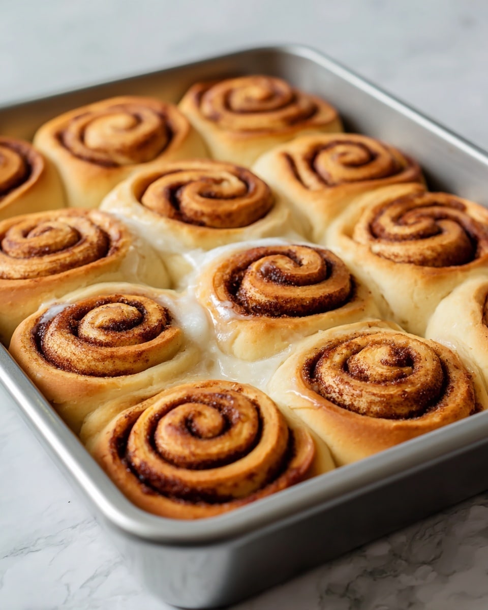 The image shows a close-up of nine cinnamon rolls arranged in a 3x3 grid inside a shiny metal baking pan. Each cinnamon roll has a golden-brown spiraled top with a dark cinnamon layer in the swirl. The dough is soft and light tan, with some areas slightly shiny from glaze or butter. The rolls are slightly touching each other, filling the pan fully on a white marbled surface. The photo was taken with an iphone --ar 4:5 --v 7