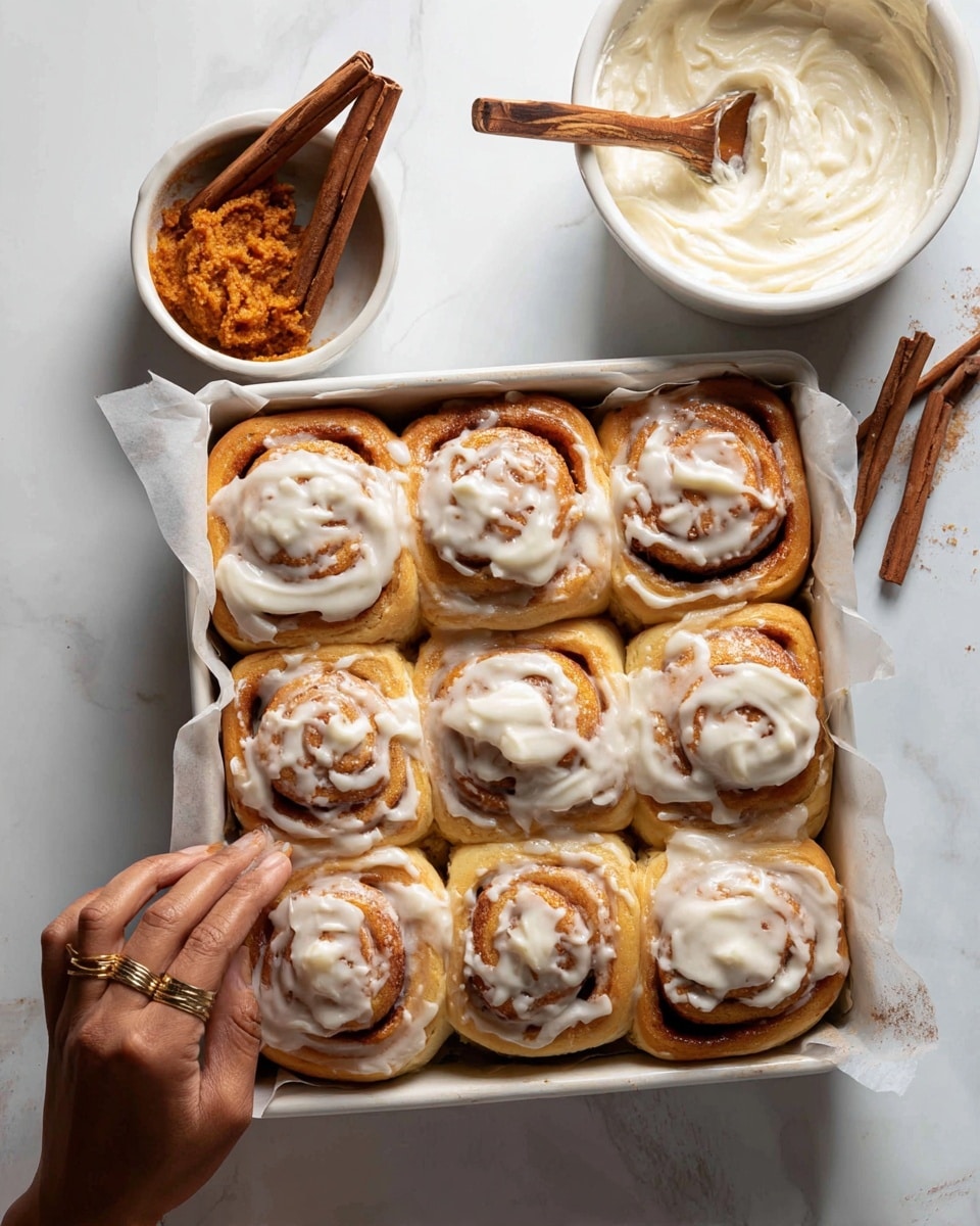 A white baking tray lined with parchment paper holds a dozen cinnamon rolls arranged in three rows and four columns, each roll showing layers of golden brown dough spiraled around a light brown cinnamon filling. The rolls are topped with a creamy white icing, spread unevenly and melting slightly into the swirls of the dough, giving a soft, smooth texture. On the top left side of the image, a small white bowl contains orange-brown cinnamon paste with several cinnamon sticks resting on top. Next to it, there is a white bowl filled with thick white icing and a wooden spoon standing inside. A woman's hand with a gold ring touches one of the cinnamon rolls on the top right corner. The background is a white marbled surface. photo taken with an iphone --ar 4:5 --v 7