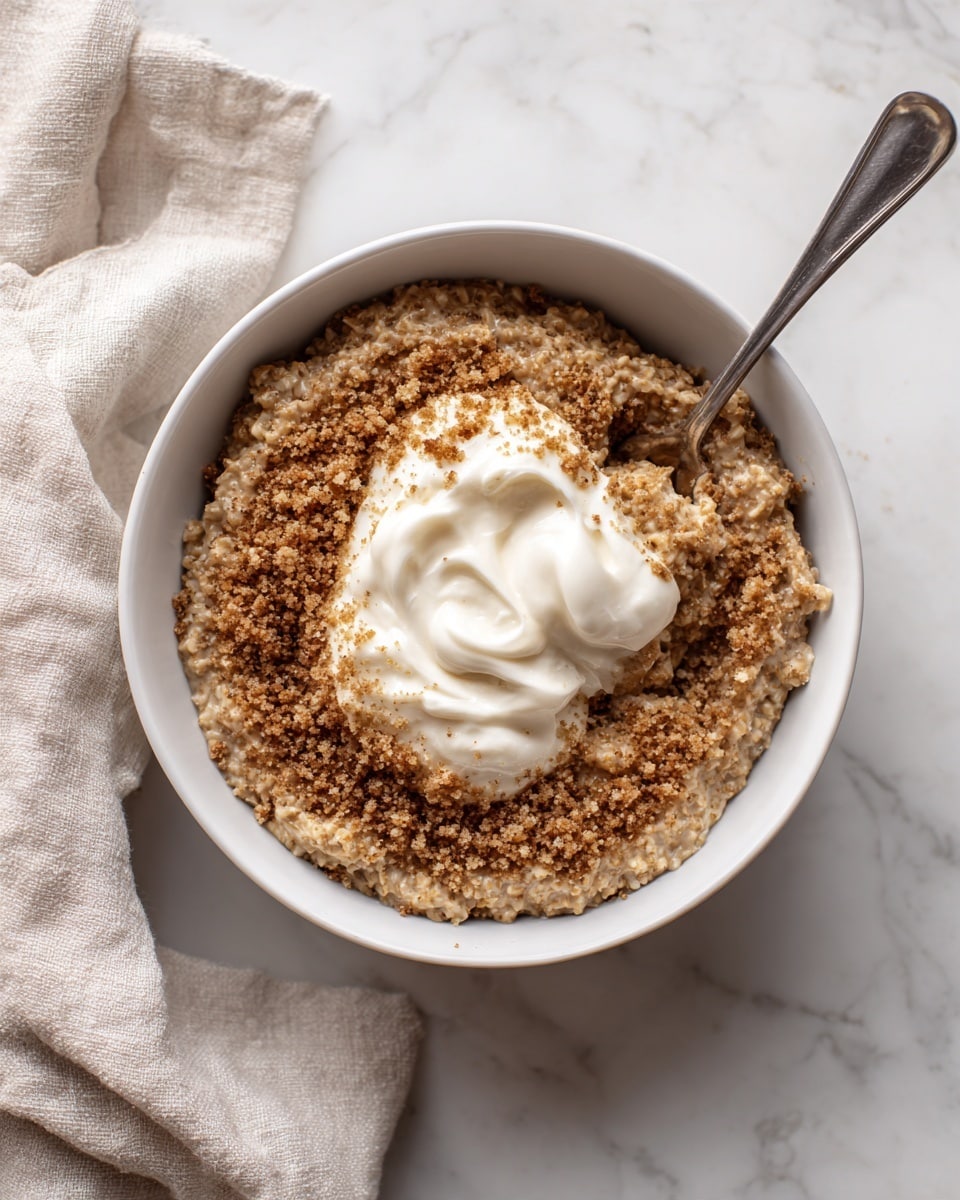 A white bowl filled with thick brown oatmeal that has a rough texture. On top, there is a layer of crushed light brown crumbs spread around the center. In the middle of the oatmeal, there is a dollop of white cream with a soft and fluffy texture. A metal spoon is placed inside the bowl, resting on the side. The bowl is set on a white marbled surface with a light beige cloth nearby. Photo taken with an iphone --ar 4:5 --v 7