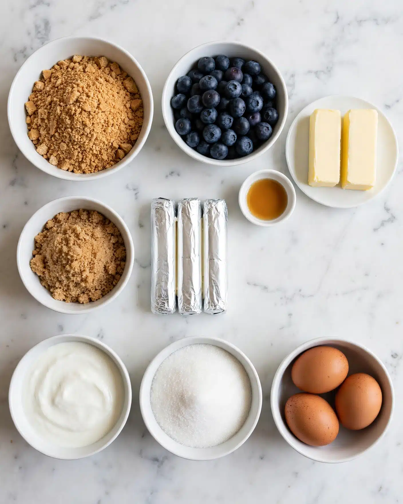 Sherry's Blueberry Cream Cheese Crumb Cake Recipe 4 The image shows nine white bowls and plates on a white marbled surface, each holding different ingredients. Starting from the top left, there is a bowl full of light brown crushed graham crackers; below it a smaller bowl with light brown sugar; below that a bowl of smooth white yogurt; next to the yogurt bowl is a bowl of granulated white sugar. To the right of the sugar bowl, there are three silver sticks of cream cheese wrapped in foil. Above these is a white bowl filled with many dark blue blueberries. At the top right, a small white plate with two sticks of creamy yellow butter is shown. Below the butter plate is a tiny white bowl holding light amber vanilla extract. Finally, at the bottom right is a white bowl containing three brown eggs. All items are neatly arranged on the smooth white marbled surface. photo taken with an iphone --ar 4:5 --v 7