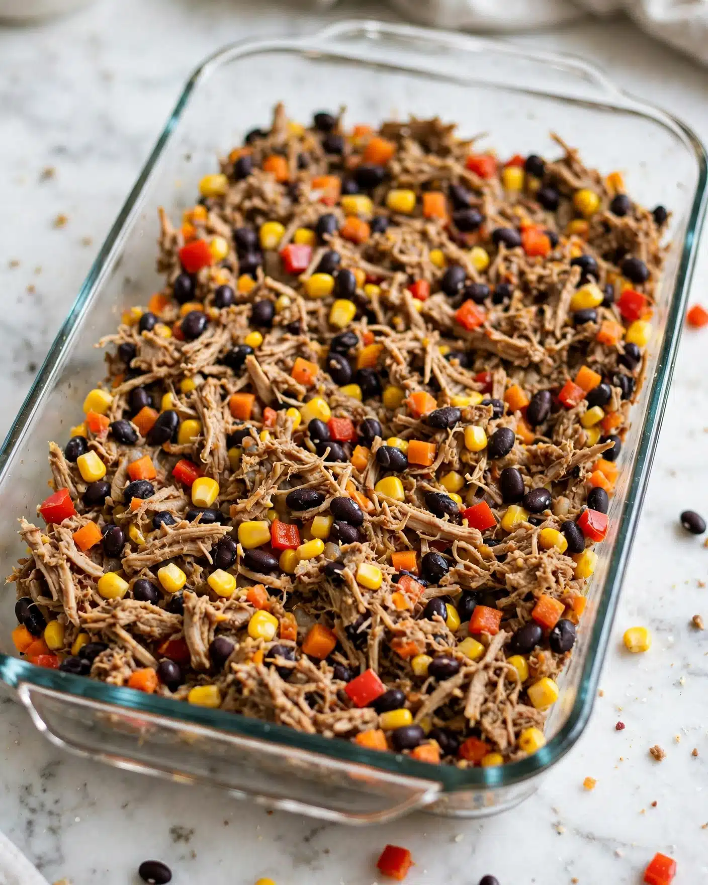 A clear glass rectangular baking dish filled with a mixed layer of shredded light brown meat, black beans, bright yellow corn, and small red and orange diced vegetables evenly spread throughout. The colorful mixture creates a textured, chunky surface with a natural, rustic appearance. The dish is set on a white marbled surface. Photo taken with an iphone --ar 4:5 --v 7