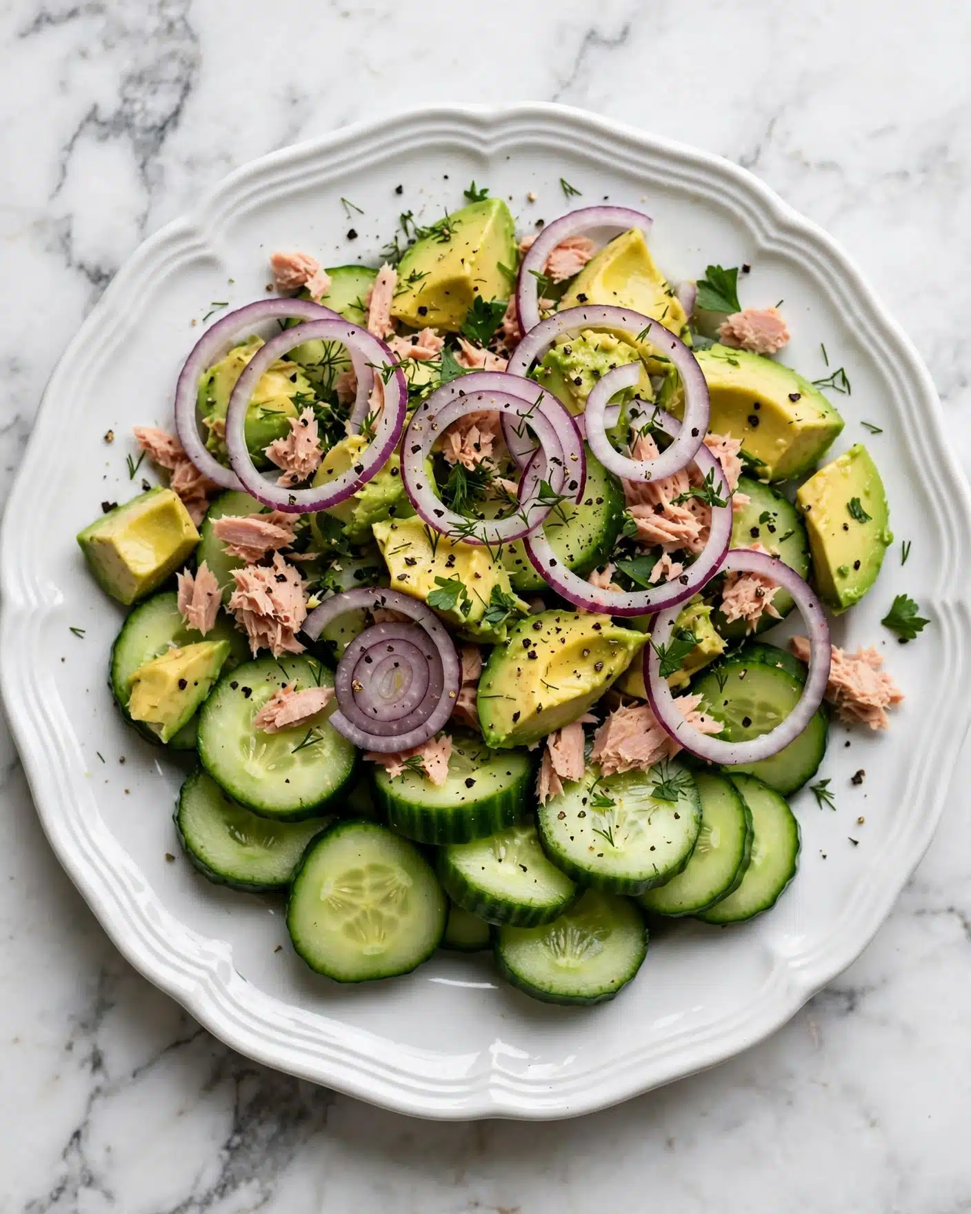 The dish is a fresh salad served in a white plate with ridged edges on a white marbled surface. It has thick slices of green cucumber layered mostly at the bottom and mixed throughout. There are chunks of yellowish-green avocado scattered across the plate, giving a smooth and creamy texture. Thin rings of purple-red onion are spread over the salad, adding a pop of color and slight crunch. Light pink shredded tuna is mixed evenly in small clumps throughout the salad. Sprinkled over everything are small green herb pieces, adding a fresh and leafy look. The salad is lightly sprinkled with black pepper for a slight speckled detail. photo taken with an iphone --ar 4:5 --v 7