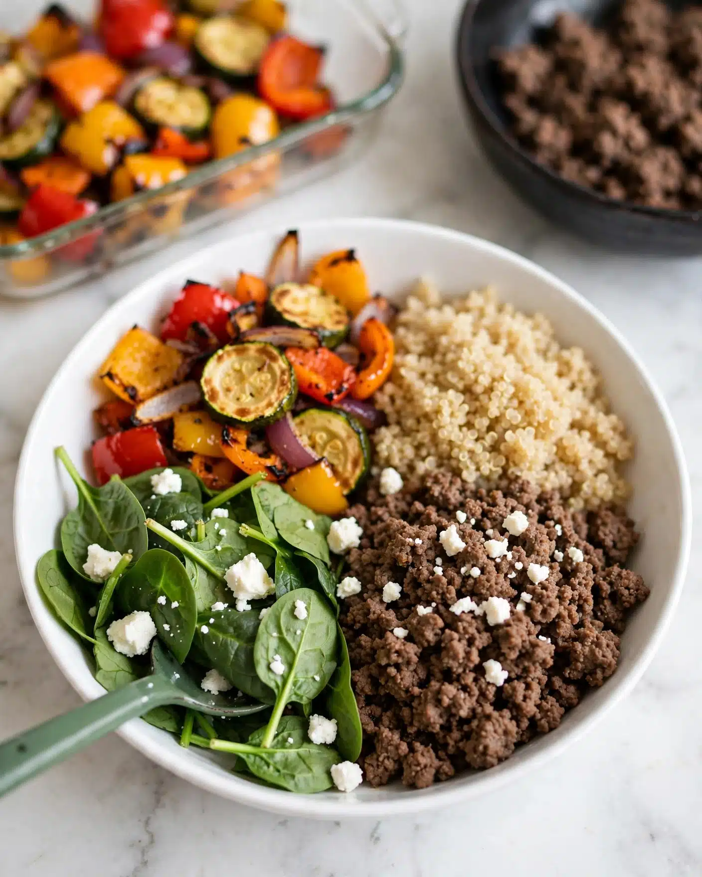 A white bowl holds four main sections of food on a white marbled surface. The first section at the bottom left contains fresh green spinach leaves, some sprinkled with small white cheese crumbles. Next to it, at the bottom right, is a mound of cooked ground beef, dark brown in color with more white cheese crumbles scattered on top. At the top right, there is a serving of light beige cooked grains resembling quinoa. The top left section has bright roasted vegetables including red, yellow, and orange bell peppers with zucchini slices, all mixed with thin slices of red onion. A green spoon is partially resting in the spinach. In the background, a glass dish of more roasted vegetables and a dark bowl of additional ground beef are slightly out of focus. Photo taken with an iphone --ar 4:5 --v 7