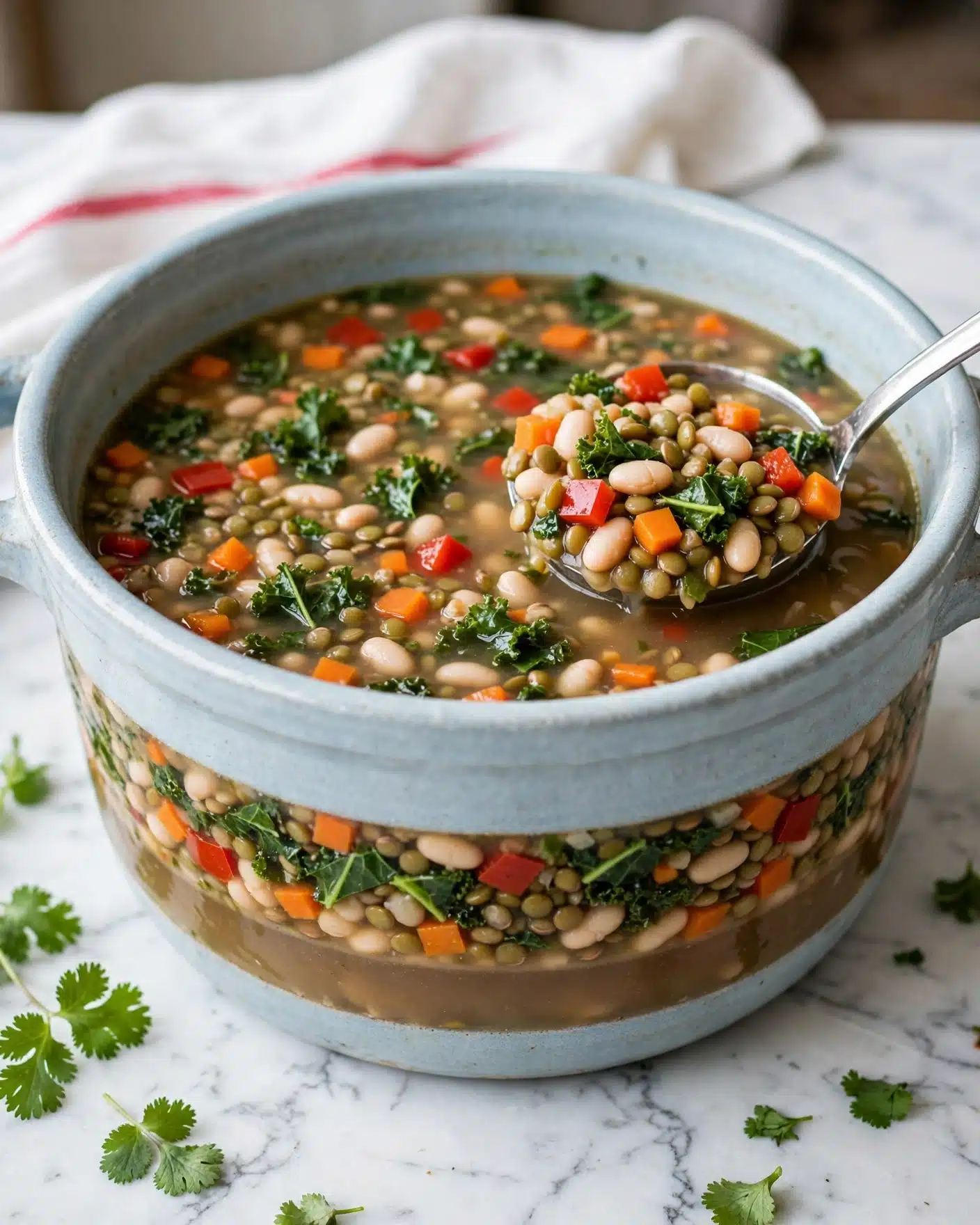 A large light blue pot sits on a white marbled surface, filled with thick soup showing three visible layers: a clear broth at the bottom with a light brown color, a mid-layer packed with small chopped orange carrots, red bell peppers, green kale leaves, white beans, and green lentils, and a floating layer of these ingredients mixed evenly with the broth on top. A silver metal ladle rests inside the pot, scooping up a portion of the soup that clearly shows the mix of colors and textures. Small green leaves of fresh cilantro are placed around the pot, and a white cloth with a thin red stripe is in the background. Photo taken with an iphone --ar 4:5 --v 7