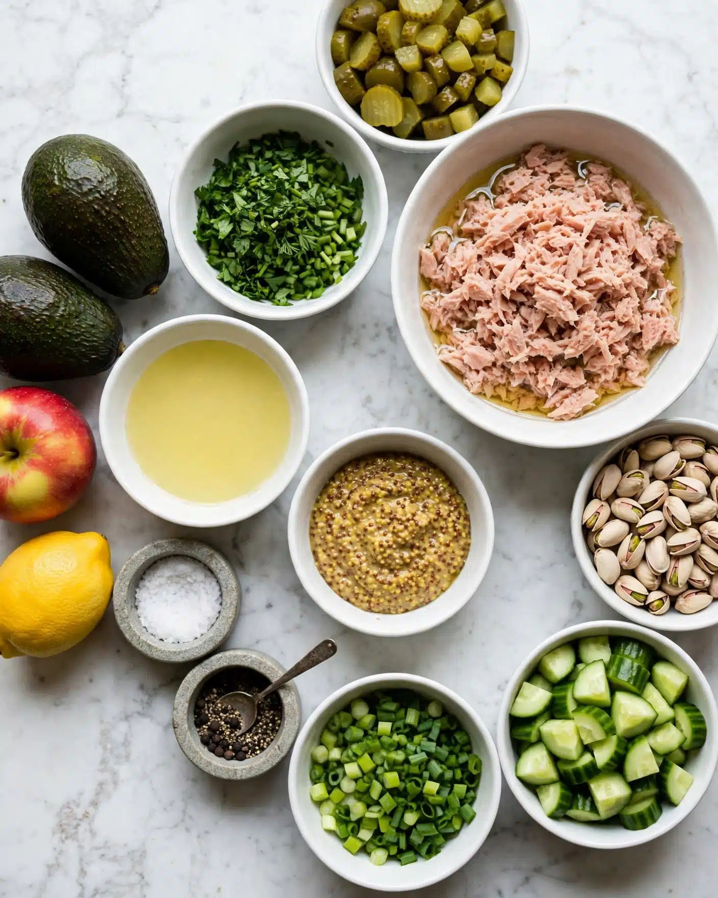 A top-down view of various ingredients laid out on a pale blue wooden surface changed to a white marbled texture, featuring a white bowl filled with light pink shredded tuna in light oil on the right. Around it are multiple white bowls each with chopped pickles, whole pistachios, chopped cucumbers, chopped green onions, grainy mustard, light yellow liquid, and chopped fresh green herbs. On the left side are two whole dark green avocados, a red and yellow apple, and a bright yellow lemon. There are two small round stone containers, one with coarse salt and the other with black pepper and a small spoon inside. The image captures the fresh and raw look of the ingredients perfectly, photo taken with an iphone --ar 4:5 --v 7