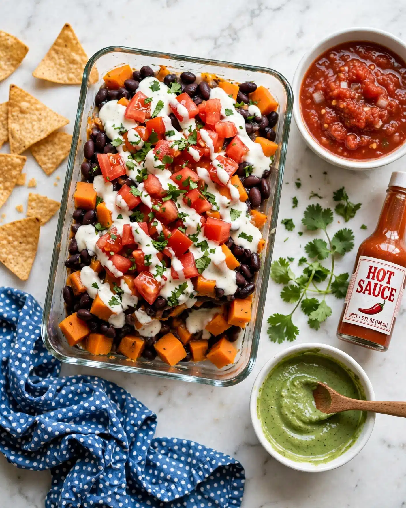 A clear rectangular glass dish filled with a layered casserole featuring a base of cooked black beans and diced orange sweet potatoes, topped with red tomato chunks and finely chopped green herbs scattered across the surface. White creamy sauce is drizzled unevenly over the top, adding contrast to the vibrant warm colors. The dish is placed on a white marbled surface next to a white bowl filled with chunky red salsa, another white bowl with a green creamy sauce and a wooden spoon inside, a bottle of hot sauce, fresh cilantro sprigs, and scattered tortilla chips on the left side. A blue cloth napkin with white spots is crumpled near the bottom left corner. Photo taken with an iphone --ar 4:5 --v 7