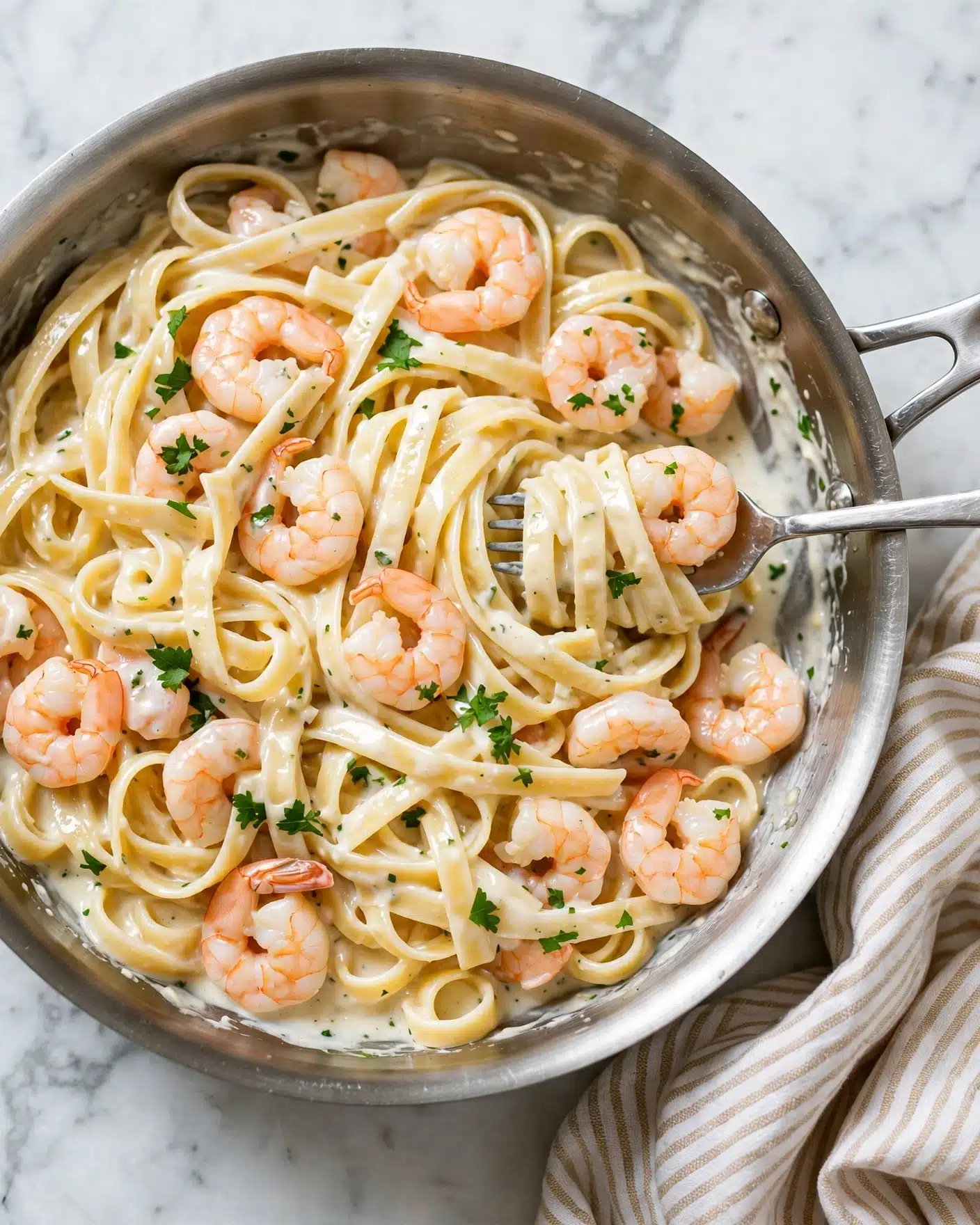 A silver pan filled with creamy white fettuccine pasta layered with a mix of light pink shrimp scattered evenly throughout. The shrimp have a slight orange tint and look tender, sitting on top and mixed well into the pasta layers. The fettuccine noodles are wide, soft, and coated with a smooth, pale creamy sauce giving them a shiny texture. Small green parsley pieces are sprinkled on top and mixed in lightly for a fresh contrast. A silver fork is placed inside the pan, slightly lifting some pasta and shrimp. The pan rests on a white marbled surface with a soft striped cloth nearby. Photo taken with an iphone --ar 4:5 --v 7