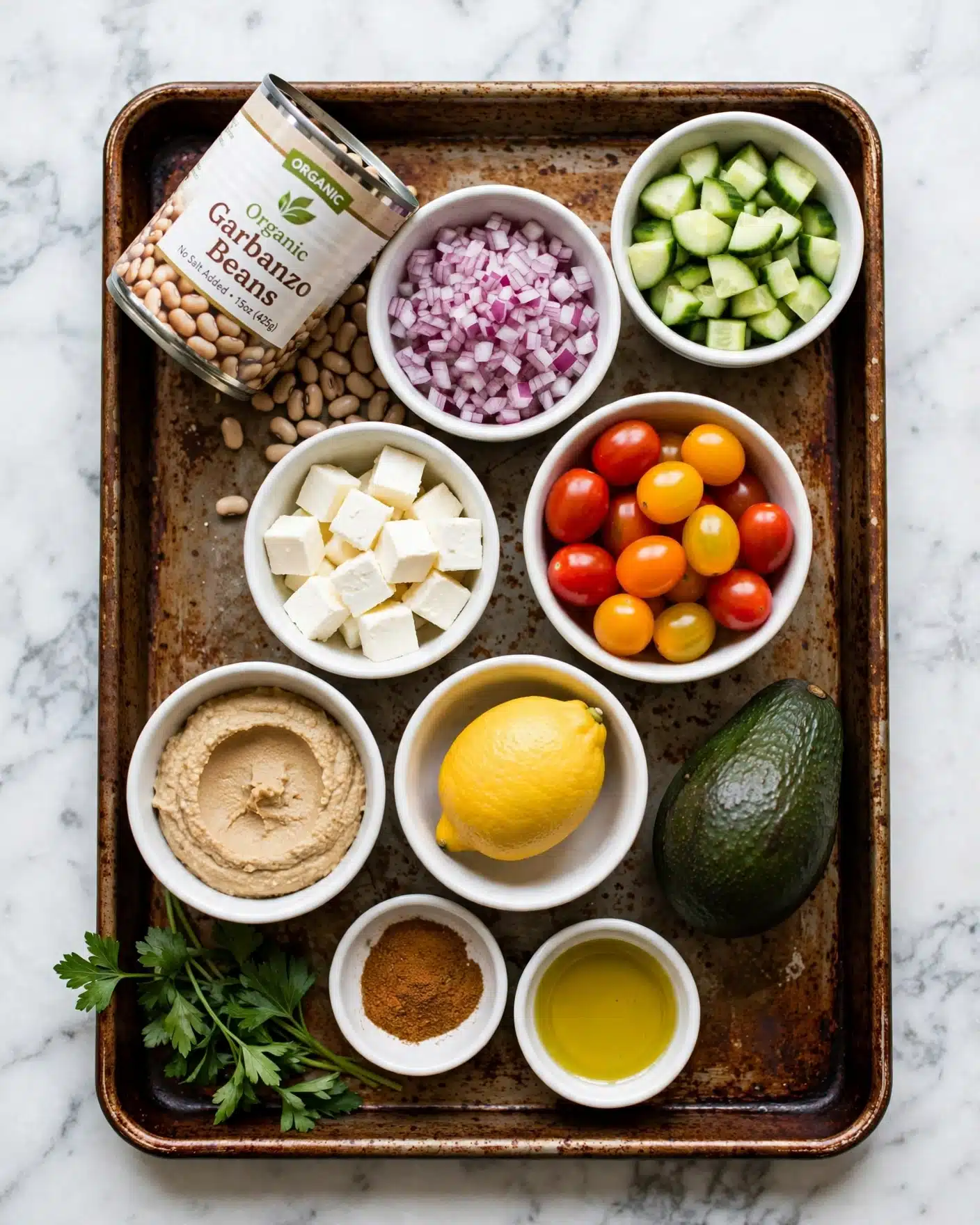 Corn, Tomato, and Avocado Chickpea Salad Recipe 4 A rusty brown baking tray holds various white bowls and ingredients arranged neatly on a white marbled surface. At the top left, a tilted can labeled