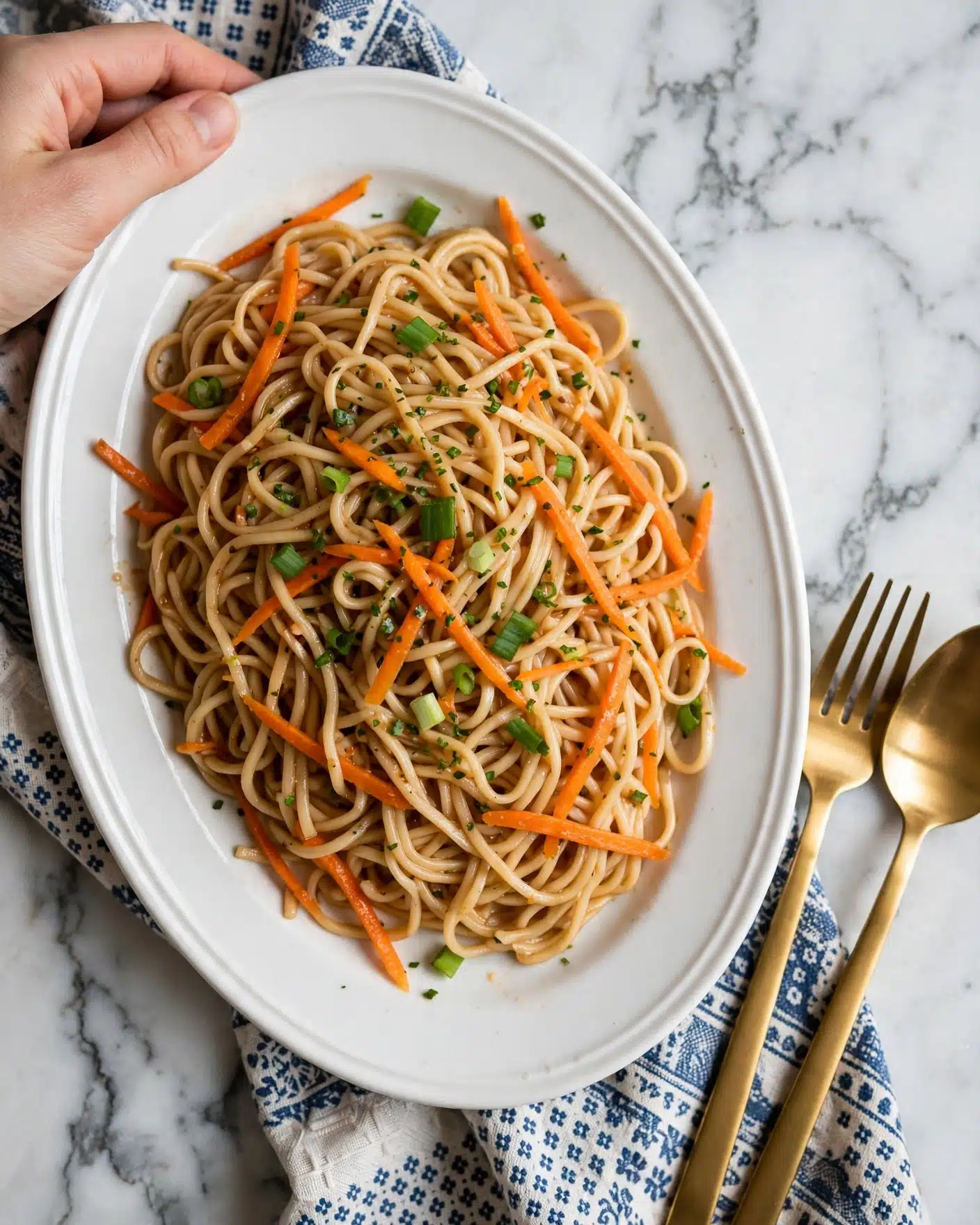 Summer Veggie Stir-Fry with Noodles Recipe 5 The image shows a white oval plate filled with a colorful noodle dish. The noodles have a light brown color, mixed with thin orange strips of carrot and small green pieces of chopped green onions scattered on top. The texture of the noodles looks soft and lightly coated with sauce. The plate is placed on a white marbled surface with a patterned cloth underneath. A gold fork and spoon are placed on the right side of the plate, and a woman’s hand is gently holding the edge of the plate in the top left corner. photo taken with an iphone --ar 4:5 --v 7