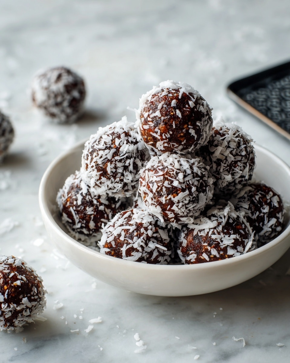 A white bowl filled with round, dark brown balls covered partly with white shredded coconut. The balls are stacked in a small pile, with some balls fully covered in coconut and others only partly. The bowl sits on a white marbled surface with a few more balls scattered around it. Nearby is a black rectangular object with a shiny, patterned texture. The light is soft and natural, highlighting the rough texture of the balls and the contrast between the dark brown and white coconut. photo taken with an iphone --ar 4:5 --v 7