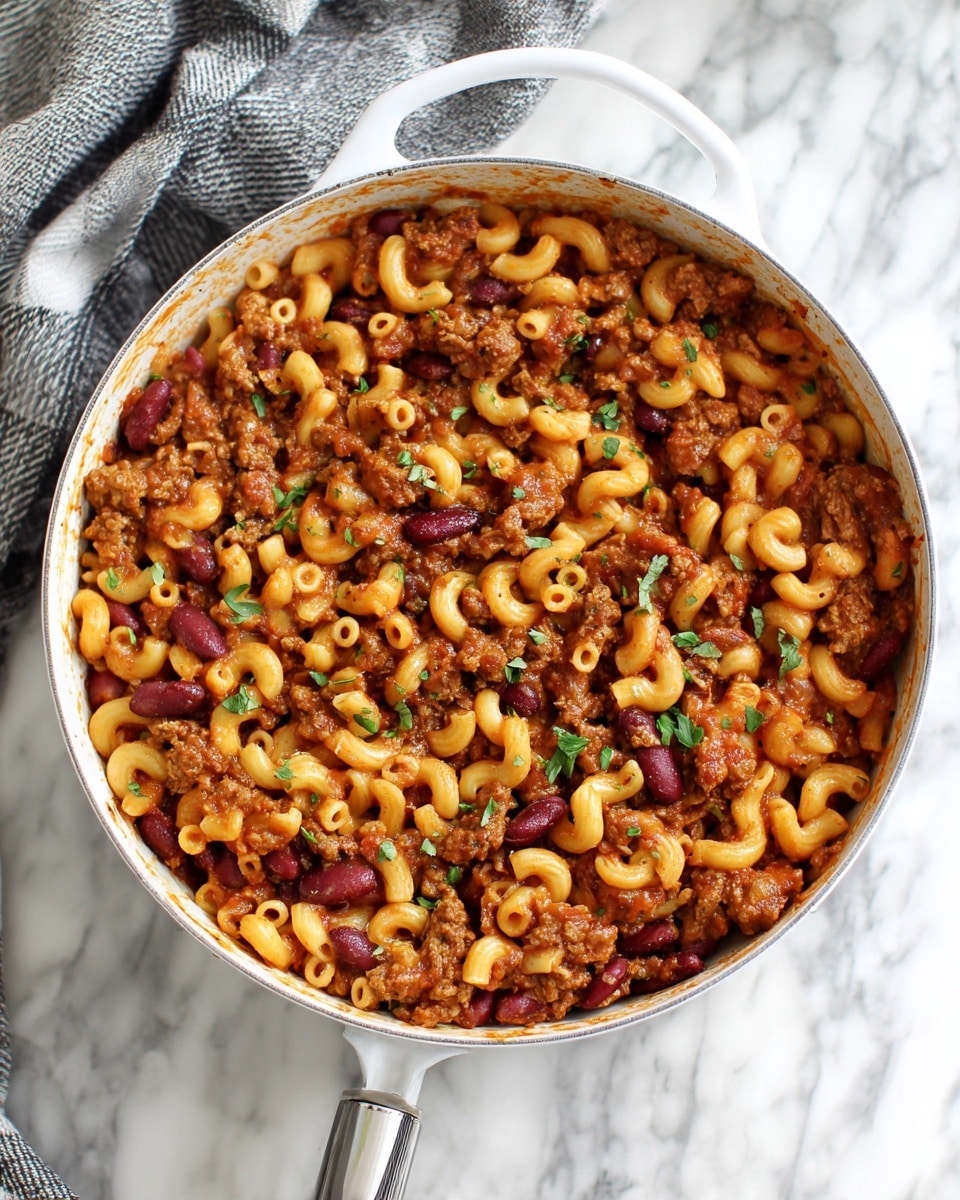 The image shows a white deep pan filled with a thick mix of small elbow macaroni, red kidney beans, and ground meat well cooked in a tomato-based sauce. The dish is scattered with small pieces of green herbs on top, adding a touch of freshness. The texture appears rich and slightly saucy, with the meat and beans evenly spread throughout the pasta. The pan handle is visible at the bottom, and the pan sits on a white marbled surface with a gray and white checkered cloth nearby. photo taken with an iphone --ar 4:5 --v 7