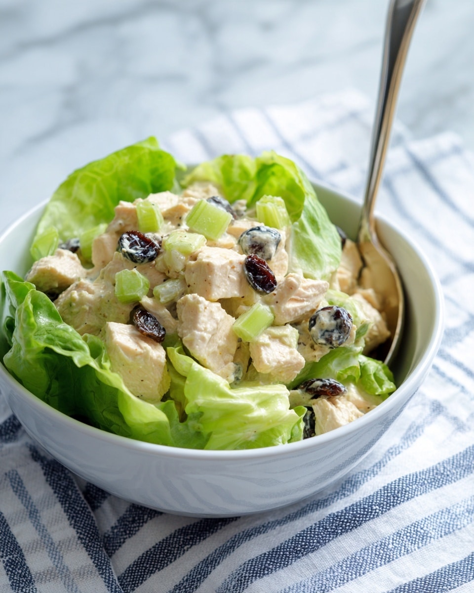 A white bowl filled with a creamy salad that has visible pieces of light tan chicken, green celery chunks, and dark raisin-like bits mixed throughout. The salad sits on a bed of bright green lettuce leaves that line the inside of the bowl. A shiny silver spoon rests on the right edge of the bowl. The bowl is placed on a white and blue striped cloth over a white marbled surface, creating a fresh and clean look. Photo taken with an iphone --ar 4:5 --v 7