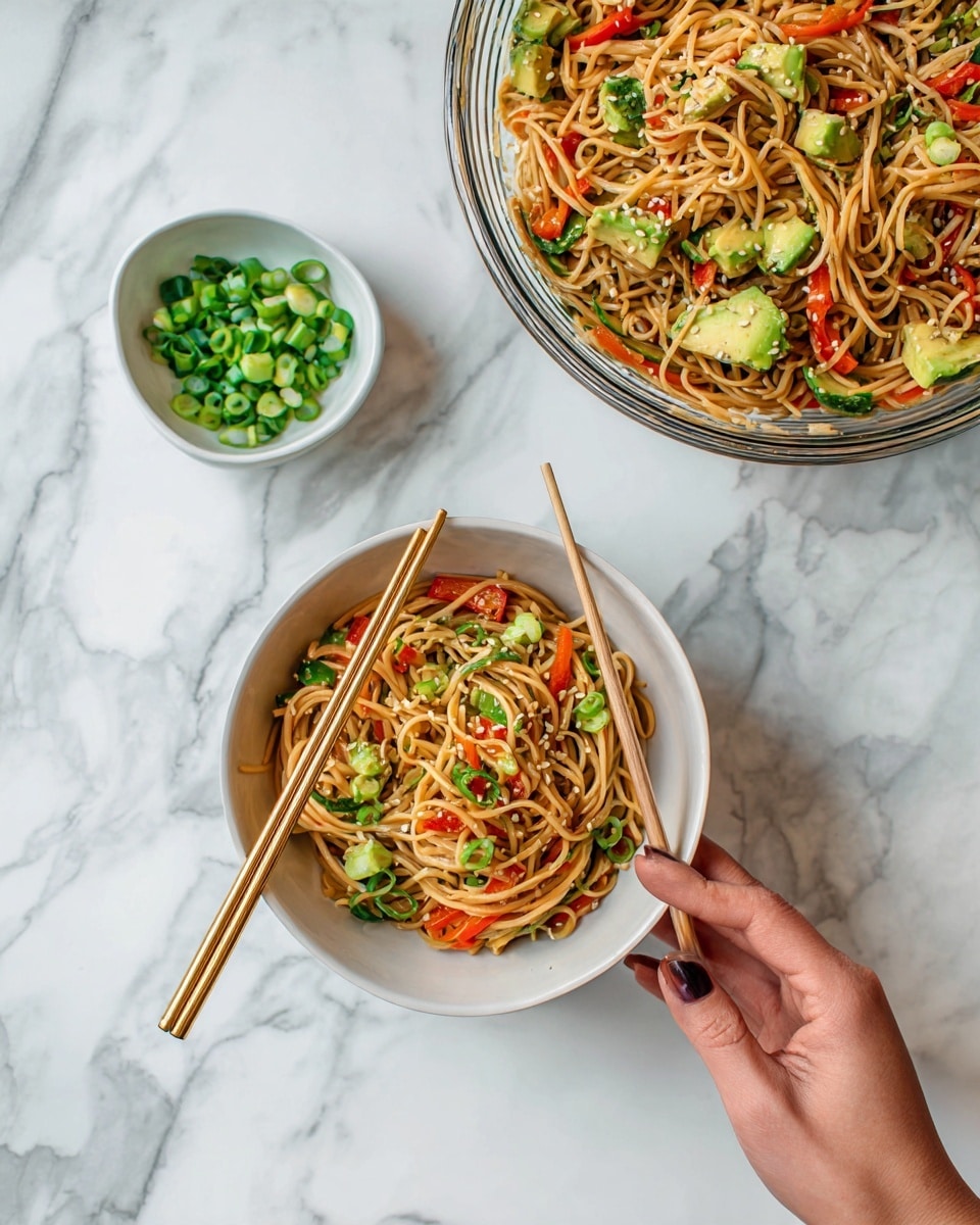The image shows a white bowl filled with light brown noodles mixed with small pieces of red bell pepper and green vegetables. A pair of chopsticks rests inside the bowl. Above it, a larger clear glass bowl contains more of the same noodle salad with visible chunks of avocado on top. To the right, a small white bowl holds chopped green onions. The scene is set on a white marbled surface, and a woman's hand holds one of the golden chopsticks in the bottom left corner. Photo taken with an iphone --ar 4:5 --v 7