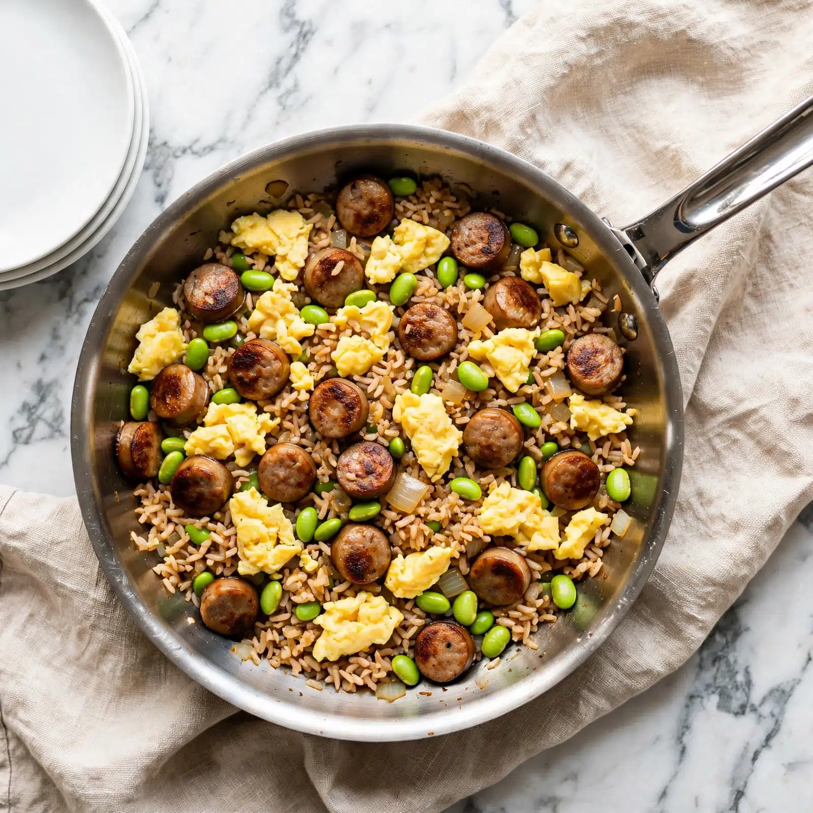 square image A skillet filled with a colorful fried rice dish shows a mix of fluffy yellow scrambled egg pieces scattered through medium brown fried rice. Small round sausage slices with a darker browned surface are spread evenly throughout the pan along with bright green edamame beans adding pops of color. There are also small bits of cooked onion mixed in, contributing a light translucent texture. The skillet is silver with a shiny metal handle, sitting on a beige cloth. White plates are stacked nearby on a white marbled surface. photo taken with an iphone --ar 4:5 --v 7