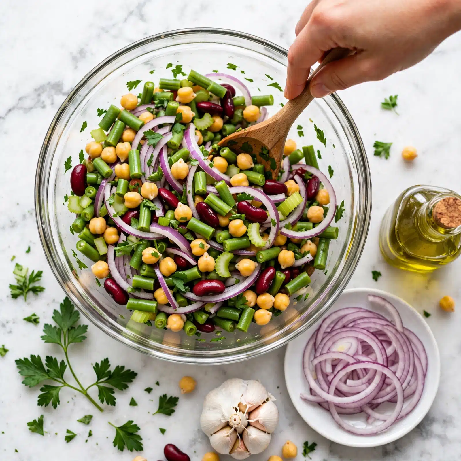 square image A clear glass bowl full of a colorful salad with layers of green beans cut into small pieces, yellow chickpeas, dark red kidney beans, and thin purple onion slices. There are also green celery pieces and chopped parsley scattered throughout. A woman's hand is holding a wooden spoon inside the bowl, mixing the salad. The bowl sits on a white marbled surface with parsley leaves, garlic bulb, a white plate with onion slices, and a glass bottle of oil around it. Photo taken with an iphone --ar 4:5 --v 7